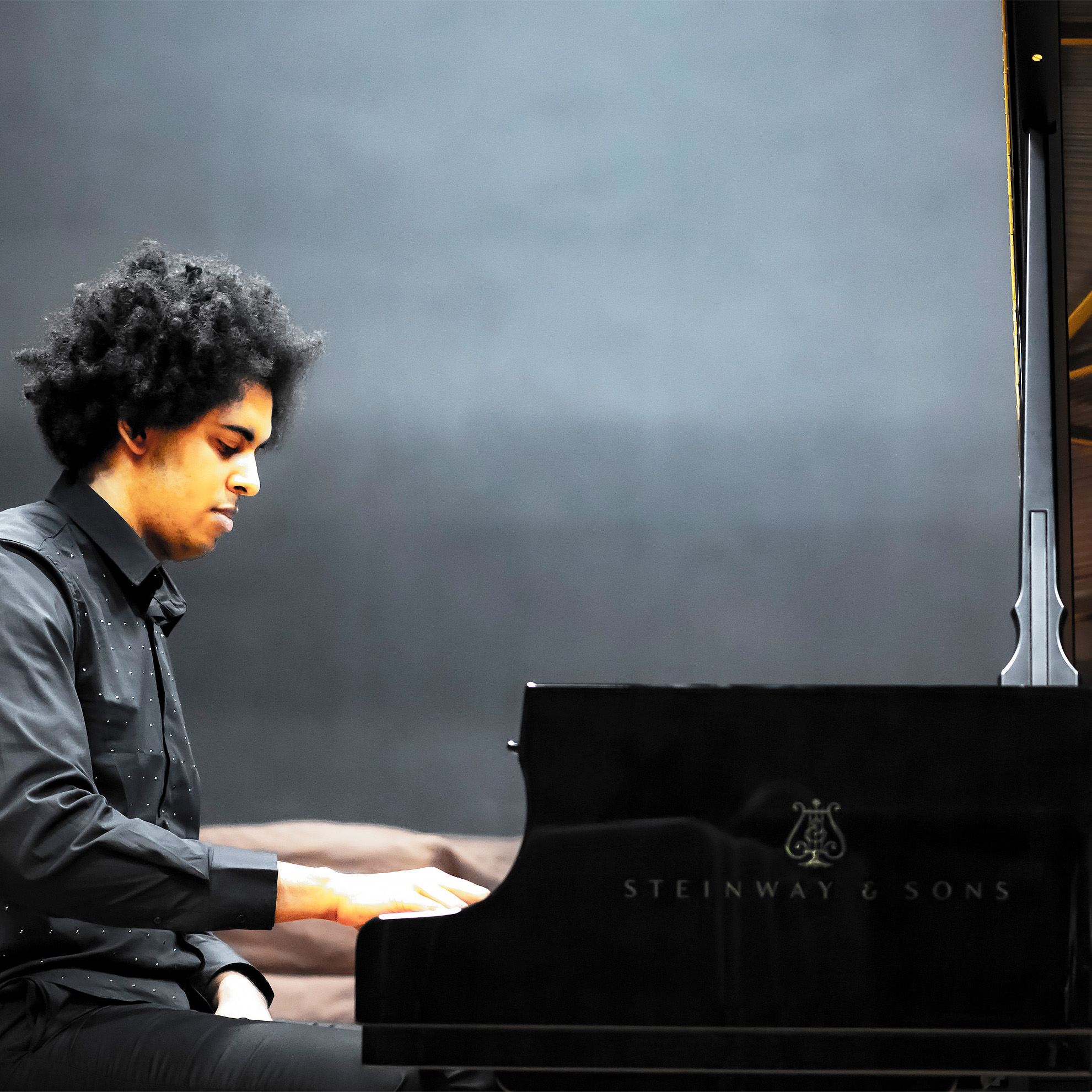 A male student playing the piano, with a dark background behind him